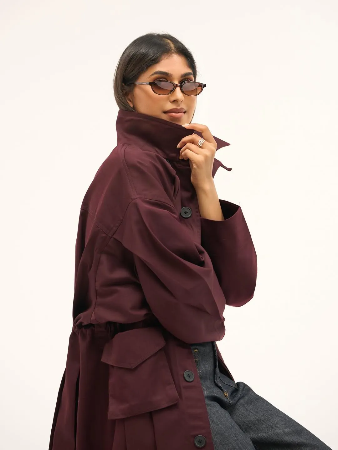 Woman in the cherry trench coat holding its raised collar, wearing oval tinted sunglasses